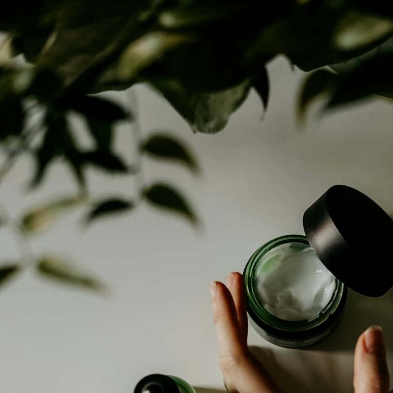 Mano sosteniendo frasco de crema de cuidado de piel junto a sérum verde, planta al fondo
